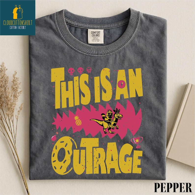 a gray t-shirt with a vibrant graphic design, including a dinosaur, flowers, and the text "This is an Outrage".