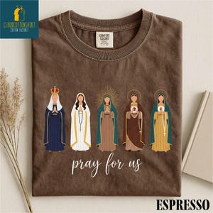 May include: Espresso-colored t-shirt featuring a graphic of five religious figures in robes and crowns. The words "pray for us" are written in cursive below the figures. The shirt has a Comfort Colors tag.