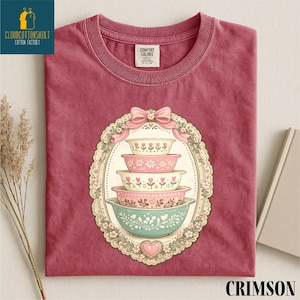May include: Crimson-colored t-shirt featuring a stack of decorative bowls in pink, white, and teal, adorned with floral and heart motifs. The design is framed by a decorative oval with a pink bow at the top. The shirt has a crew neck and short sleeves.