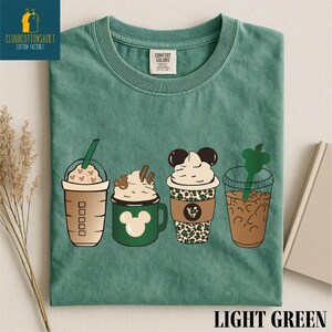 May include: Light green t-shirt featuring four illustrated coffee drinks. The drinks include a Frappuccino, a Mickey Mouse mug with whipped cream, a Mickey Mouse-themed cup with leopard print, and an iced coffee with a green straw.