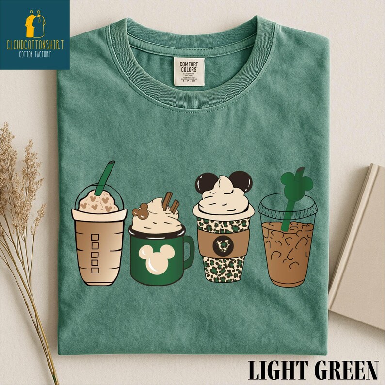 May include: Light green t-shirt featuring four illustrated coffee drinks. The drinks include a Frappuccino, a Mickey Mouse mug with whipped cream, a Mickey Mouse-themed cup with leopard print, and an iced coffee with a green straw.