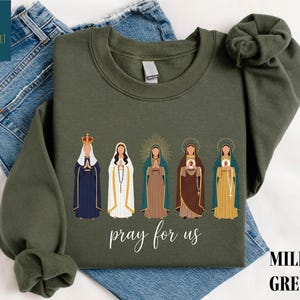 May include: Military green sweatshirt featuring an illustration of five religious figures. The figures are depicted in various colors, with the text "pray for us" in a script font. The sweatshirt is displayed on a pair of blue jeans.