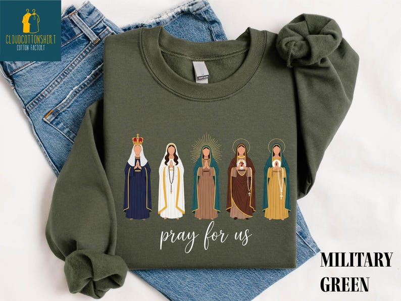 May include: Military green sweatshirt featuring an illustration of five religious figures. The figures are depicted in various colors, with the text "pray for us" in a script font. The sweatshirt is displayed on a pair of blue jeans.