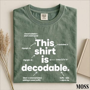 May include: A sage green t-shirt with white text that reads "This shirt is decodable." The design includes linguistic annotations with arrows pointing to specific letters and words, explaining phonics rules like digraphs, syllables, and suffixes.