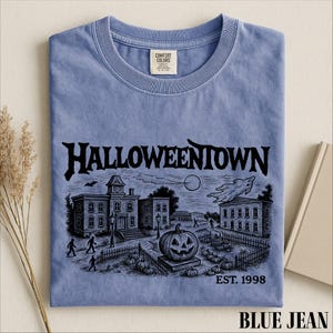 A blue t-shirt with a Halloween-themed illustration, including a pumpkin patch, buildings, and a ghost, along with the text "Halloweentown" and "EST. 1998".