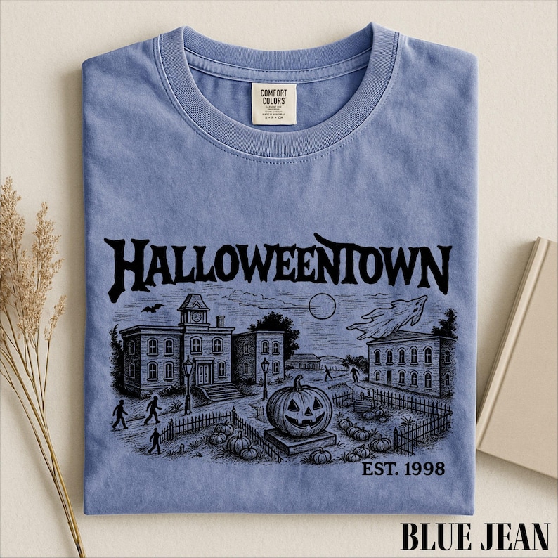 A blue t-shirt with a Halloween-themed illustration, including a pumpkin patch, buildings, and a ghost, along with the text "Halloweentown" and "EST. 1998".