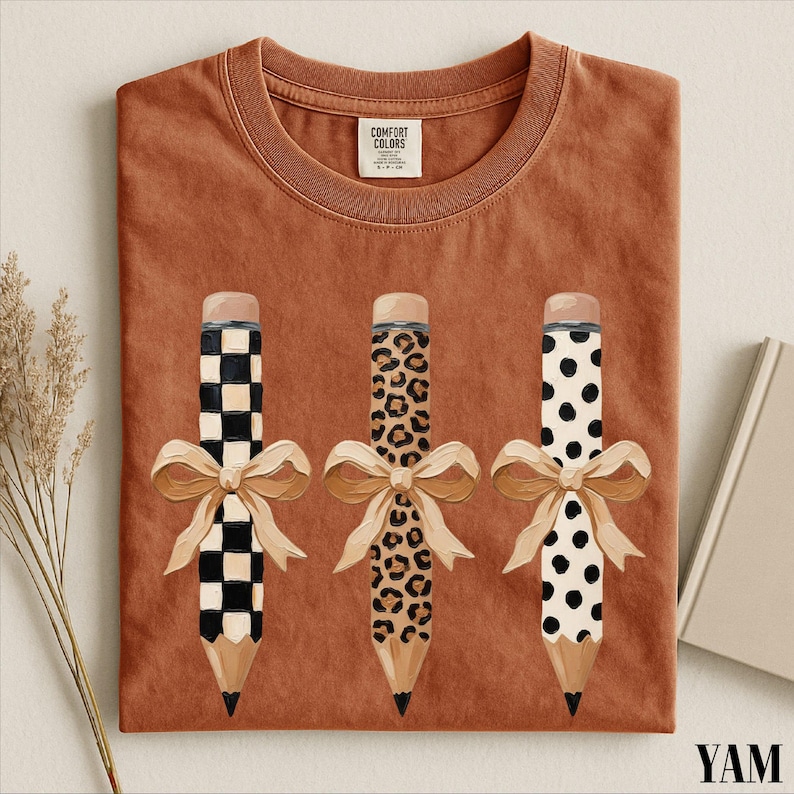 a red t-shirt with a design of three pencils with bows, each with a different pattern: a checkered pattern, a leopard print pattern, and a polka dot pattern.