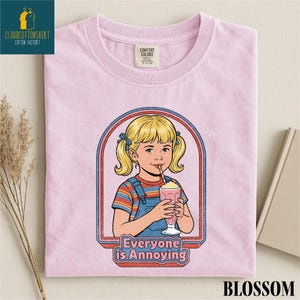 A pink t-shirt with a graphic design depicting a young girl with blonde hair and a straw, holding a milkshake, with the text "Everyone is Annoying" below the image.