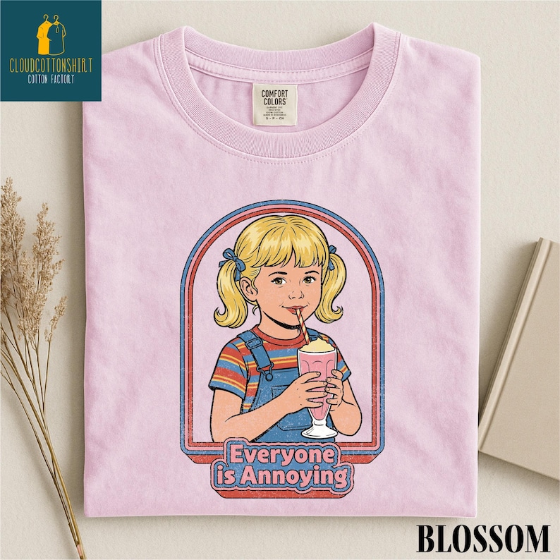 A pink t-shirt with a graphic design depicting a young girl with blonde hair and a straw, holding a milkshake, with the text "Everyone is Annoying" below the image.