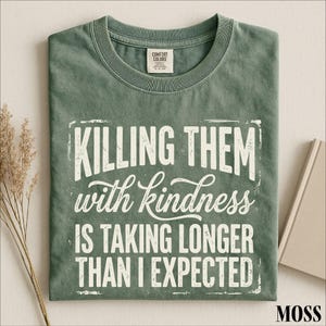 A green t-shirt with a white text design that reads "Killing them with kindness is taking longer than I expected".