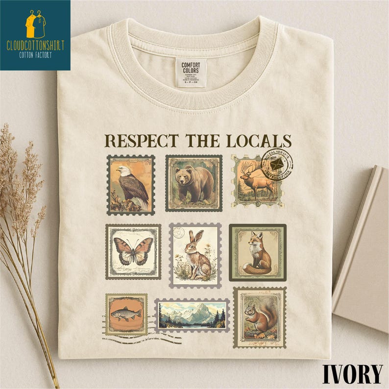 May include: Ivory-colored t-shirt with the text "Respect the Locals" and postage stamp-style illustrations of wildlife, including a bald eagle, bear, deer, butterfly, rabbit, fox, fish, and squirrel. The shirt has a Comfort Colors tag.