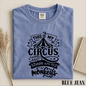May include: A blue jean colored short-sleeve t-shirt with a black graphic print. The design features a circus tent icon and the text "THIS IS MY CIRCUS AND THESE ARE MY MONKEYS" in a stylized font. The shirt has a crew neckline and a brand label.