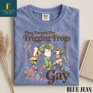 Pu&ograve; includere: T-shirt blu jeans con un design grafico con tre rane danzanti, farfalle e funghi. Il testo recita "They Turned The Frigging Frogs Gay". La maglietta ha un'etichetta Comfort Colors.