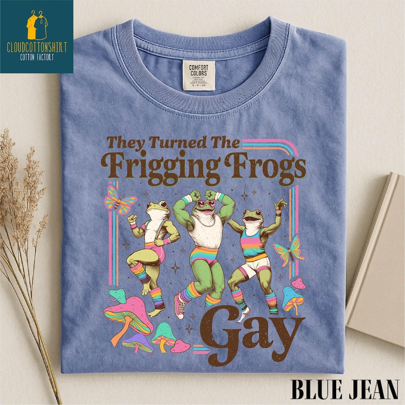 Pu&ograve; includere: T-shirt blu jeans con un design grafico con tre rane danzanti, farfalle e funghi. Il testo recita "They Turned The Frigging Frogs Gay". La maglietta ha un'etichetta Comfort Colors.