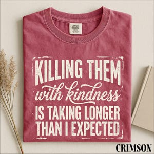 A red t-shirt with white text that reads "Killing them with kindness is taking longer than I expected" is displayed on a white background.
