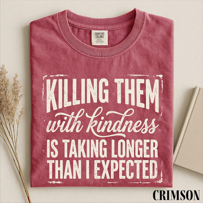 A red t-shirt with white text that reads "Killing them with kindness is taking longer than I expected" is displayed on a white background.