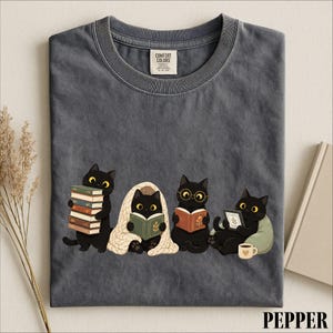 May include: A charcoal gray crew neck t-shirt featuring a graphic of four black cats. One cat holds a stack of books, one sits under a beige blanket reading, one wears round glasses while reading, and one reads a tablet next to a coffee mug.