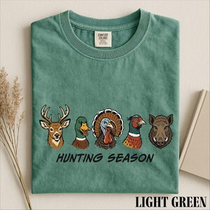 A green t-shirt with a graphic design of five different animal heads, including a deer, a turkey, a boar, a duck, and a goose, along with the text "HUNTING SEASON".