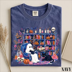 May include: A navy blue t-shirt with a graphic print featuring a white ghost reading a book in a blue armchair, a black cat, a bookshelf filled with colorful books, lit candles, pumpkins, skulls, and bats. The shirt has a Comfort Colors label.