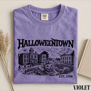 A purple t-shirt with a Halloween-themed illustration, including a pumpkin patch, a castle, and a ghost, along with the text "Halloweentown" and "EST. 1998".