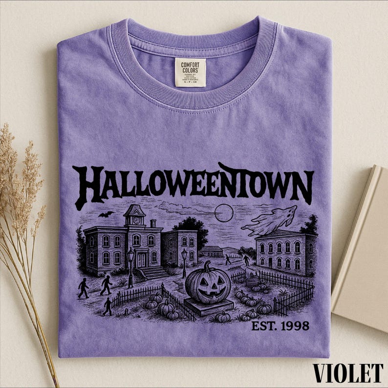 A purple t-shirt with a Halloween-themed illustration, including a pumpkin patch, a castle, and a ghost, along with the text "Halloweentown" and "EST. 1998".