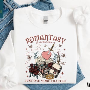 May include: White sweatshirt with a graphic design featuring the text "ROMANTASY READERS SOCIETY" and "JUST ONE MORE CHAPTER". The design includes a heart pierced by a sword, books, butterflies, roses, and a crown.