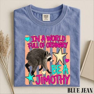 A blue t-shirt with a colorful graphic design featuring a raccoon and the text "In a world full of ordinary, be a jimothy".