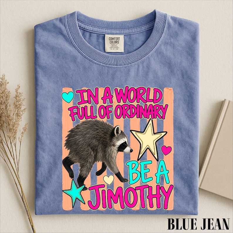 A blue t-shirt with a colorful graphic design featuring a raccoon and the text "In a world full of ordinary, be a jimothy".
