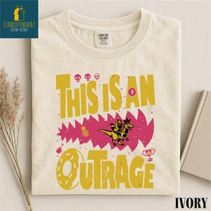 May include: Ivory-colored t-shirt with a graphic design. The design features the text "THIS IS AN OUTRAGE" in yellow, along with a pink jagged shape, a cartoon dinosaur, and other graphic elements. The shirt has a crew neck and short sleeves.