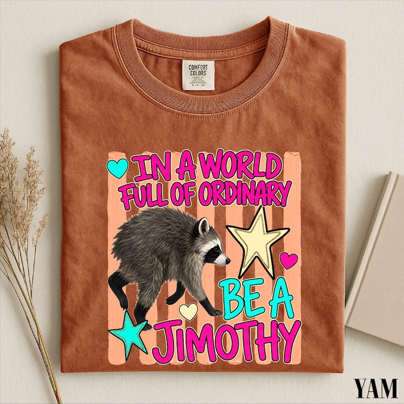 A brown t-shirt with a colorful graphic design featuring a raccoon and the text "In a world full of ordinary, be a jimothy".
