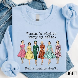 May include: A light blue crewneck sweatshirt featuring a graphic of six figures in vintage-style dresses. The text reads: Women's rights vary by state. Men's rights don't. The sweatshirt is displayed over a pair of blue denim jeans.