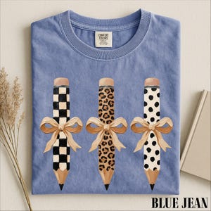 A blue t-shirt with three pencils with different designs and bows on them, along with a label and some dried flowers.