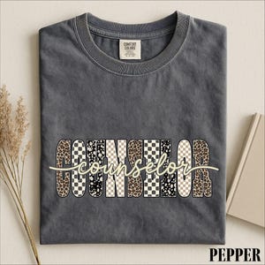 May include: A charcoal gray crew neck t-shirt with a Comfort Colors label at the inner neckline. The front features the word COUNSELOR in large block letters with varied leopard, cheetah, and checkerboard patterns, overlaid with the word counselor.