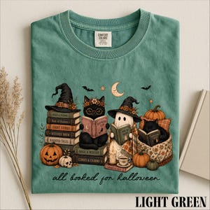 A green t-shirt with a Halloween-themed illustration of a witch, cat, ghost, pumpkin, and books, along with the text "all booked for Halloween" and a label on the collar.