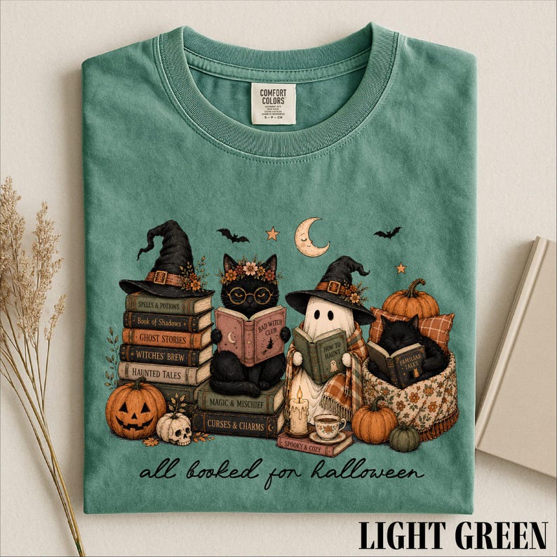 A green t-shirt with a Halloween-themed illustration of a witch, cat, ghost, pumpkin, and books, along with the text "all booked for Halloween" and a label on the collar.