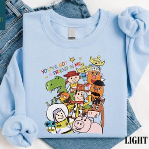K&ouml;nnte beinhalten: Hellblaues Sweatshirt mit einer bunten Grafik von Figuren aus dem Film "Toy Story". &Uuml;ber den Figuren steht der Schriftzug "YOU'VE GOT A FRIEND IN ME". Das Sweatshirt ist aus weichem Material.