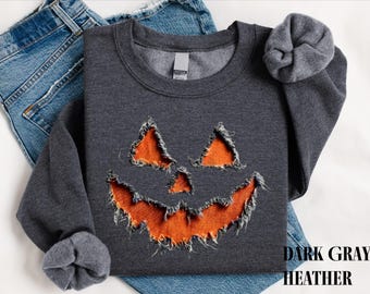 Puede incluir: Una sudadera de cuello redondo en gris oscuro jaspeado con un gráfico de cara de calabaza en naranja brillante con bordes deshilachados. El diseño muestra dos ojos triangulares, una nariz triangular pequeña y una boca ancha y dentada.