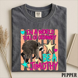 A gray t-shirt with a colorful graphic design featuring a raccoon and text that reads "In a world full of ordinary, be a jimothy".