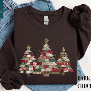May include: Dark chocolate-colored sweatshirt featuring three Christmas trees with patchwork designs in red, green, and white. Each tree is topped with a star. The text "DARK CHOCOLATE" is printed on the bottom right.