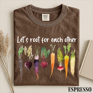 May include: A brown crew neck t-shirt featuring the white text Let's root for each other above a colorful watercolor illustration of various root vegetables including carrots, beets, and radishes in shades of purple, orange, red, and green.