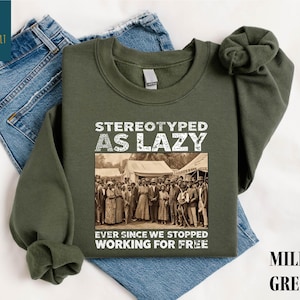 May include: Military green sweatshirt with the text "STEREOTYPED AS LAZY" above a sepia-toned image of a group of people. Below the image is the text "EVER SINCE WE STOPPED WORKING FOR FREE". The sweatshirt is displayed on top of blue jeans.