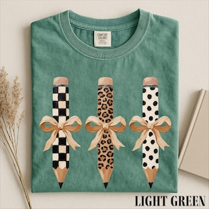 A green t-shirt with three pencils with different designs and bows on them, along with a label and some dried flowers.