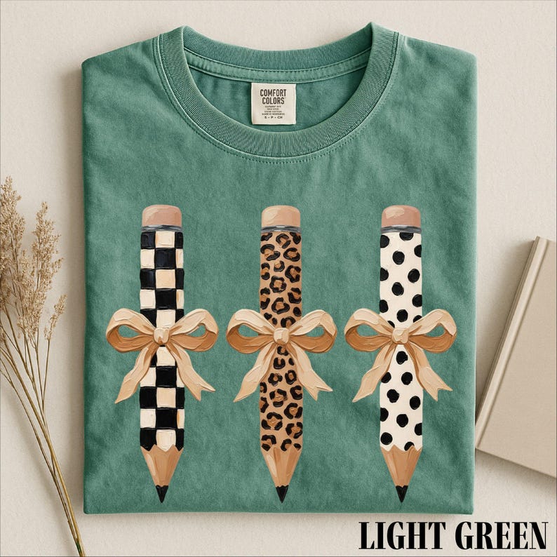 A green t-shirt with three pencils with different designs and bows on them, along with a label and some dried flowers.