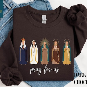 May include: Dark chocolate-colored sweatshirt featuring an illustration of five religious figures. The figures are depicted in various robes and headdresses, with the words "pray for us" in cursive script below. The words "DARK CHOCOLATE" are printed in a block font.