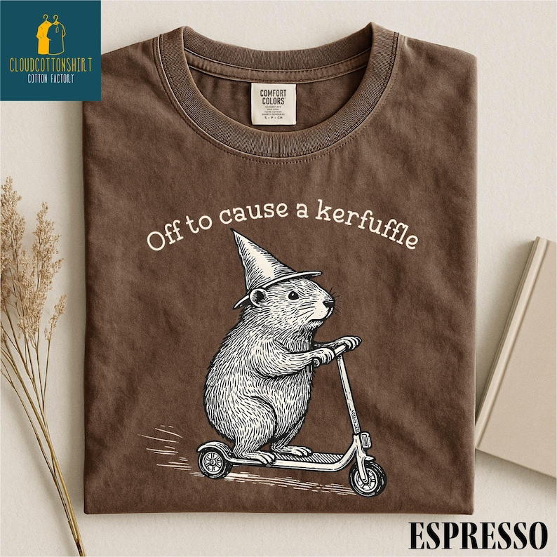 A brown t-shirt with a cartoon image of a mouse riding a scooter, wearing a pointy hat, and the text "Off to cause a kerfluffle" printed on it.