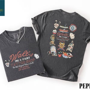 May include: Two dark gray t-shirts with a chain necklace. One shirt features the text "Walt's Ink & Paint Tattoo Shop" with various graphics. The other shirt displays a collection of tattoo-style illustrations, including a boat and a bottle.