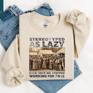 May include: Sand-colored sweatshirt with the text "STEREOTYPED AS LAZY" above a sepia-toned image of a group of people. Below the image, the text reads "EVER SINCE WE STOPPED WORKING FOR FREE". The sweatshirt is laid on top of blue jeans.