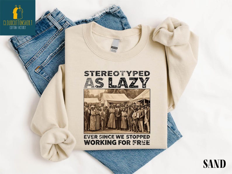 May include: Sand-colored sweatshirt with the text "STEREOTYPED AS LAZY" above a sepia-toned image of a group of people. Below the image, the text reads "EVER SINCE WE STOPPED WORKING FOR FREE". The sweatshirt is laid on top of blue jeans.