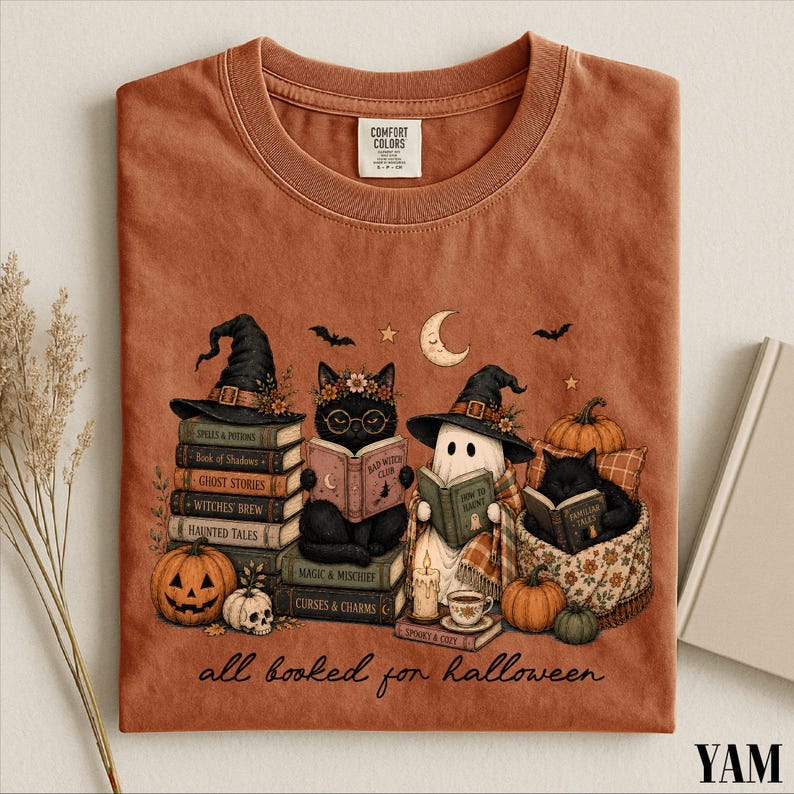 A t-shirt with a Halloween-themed graphic design featuring a witch, a cat, a ghost, and various Halloween-related items such as pumpkins, books, and a skeleton.