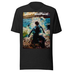 May include: Dark gray t-shirt featuring a colorful graphic print. The graphic depicts a person in a blue outfit, standing in water with waves crashing around them. The text "Lost Paintbrush" is above the figure.